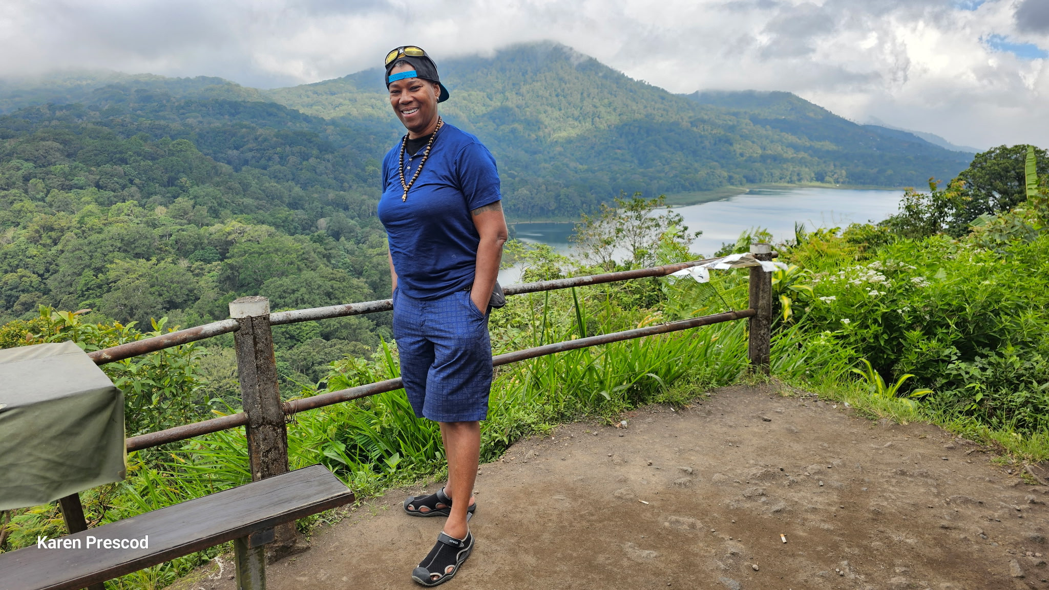 Karen smiling outdoors at a lookout beside a lake and green mountains.